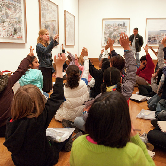 A group of students sit on the floor with their hands raised with framed art around them on the walls