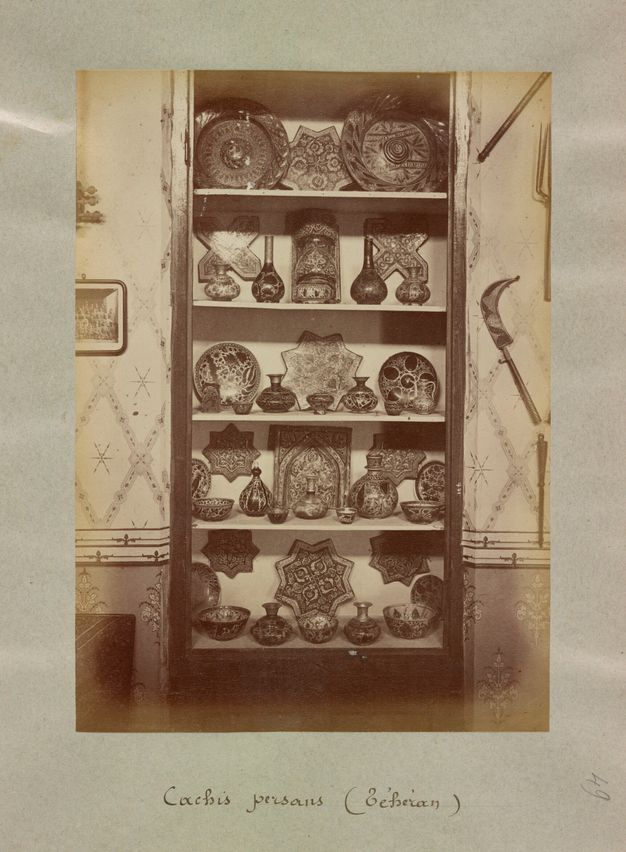 Sepia toned photograph of a cabinet against a wallpapered wall displaying luster items in an organized manner.