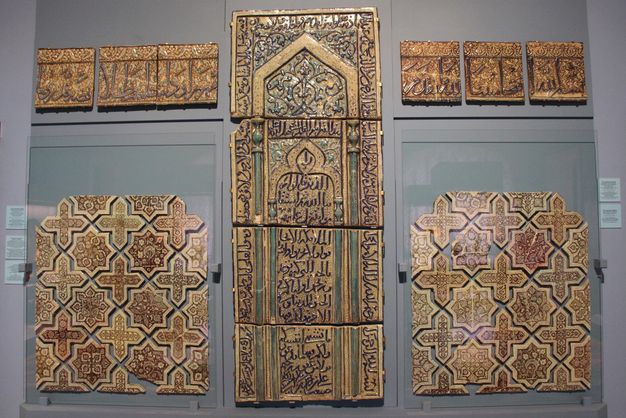 Groups of luster tiles mounted on a museum wall, including a tall middle panel, and smaller square panels with stars, crosses, and rectangles decorated with Arabic script to the upper and lower left and right.