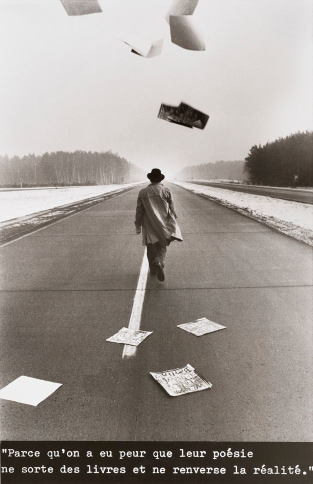 Black and white photograph of a man shown from behind walking in the center of a highway while posters flutter to the ground.