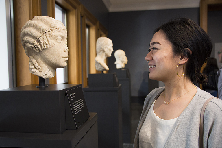 A young woman smiles at the marble bust of a young woman in a corridor at the Getty Villa.