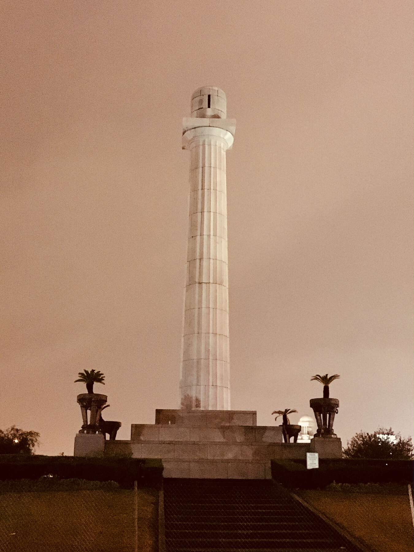 Picture at dusk of a tall white column, empty at the top; stairs and smaller pedestals ring its base.