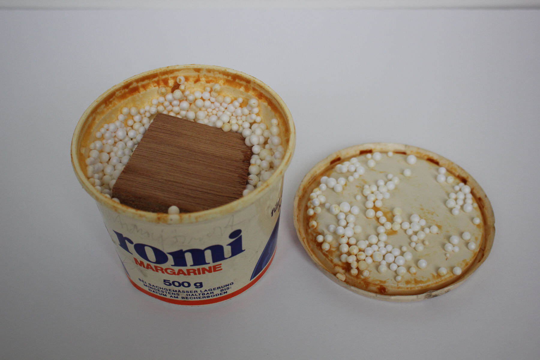 Wood submerged in ‘romi margarine’ cup full of styrafoam balls and orange discolorations with lid to the side