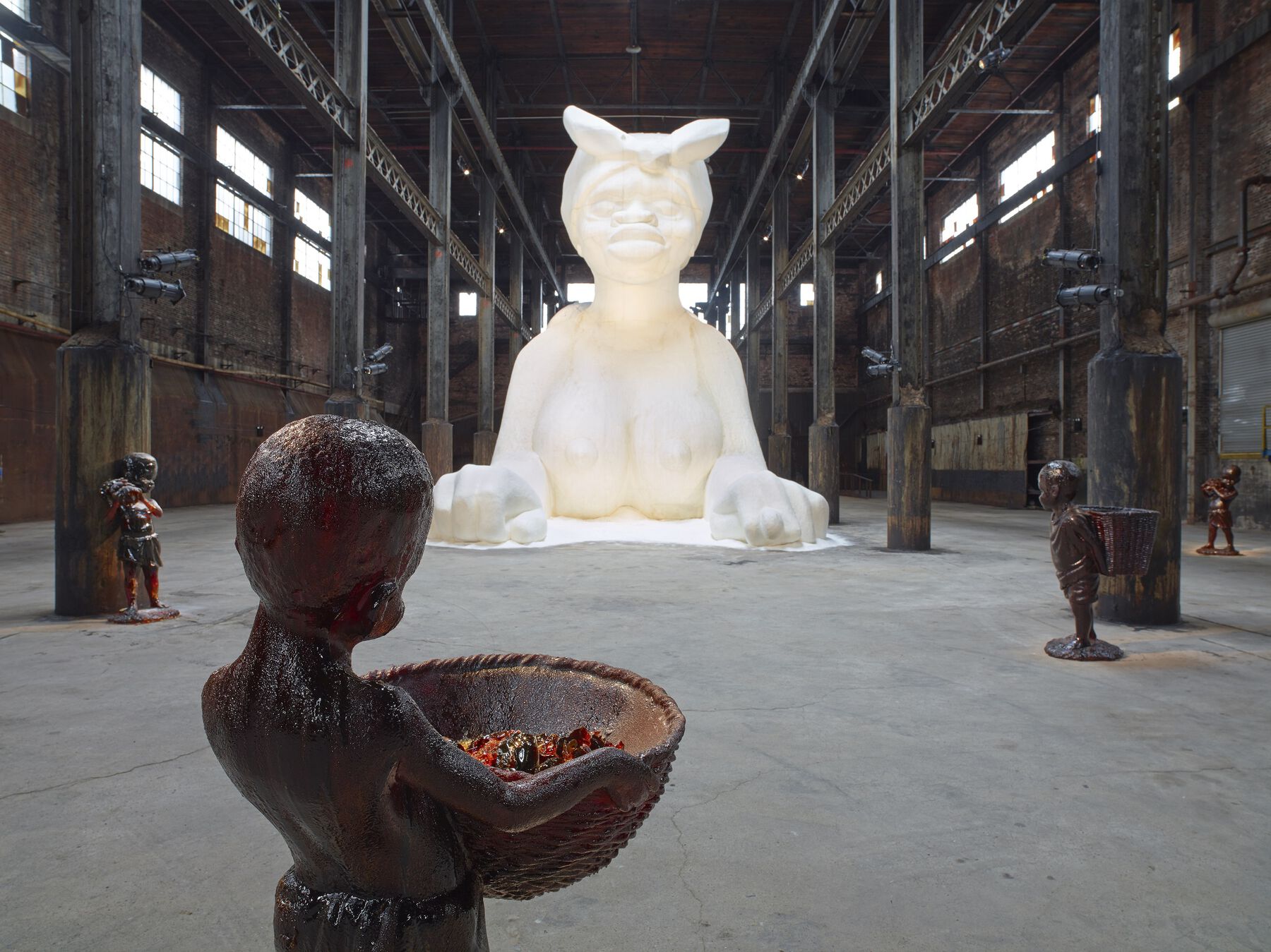 A 35-foot tall sculpture made from sugar displayed in a warehouse with small children figure sculptures scattered throughout