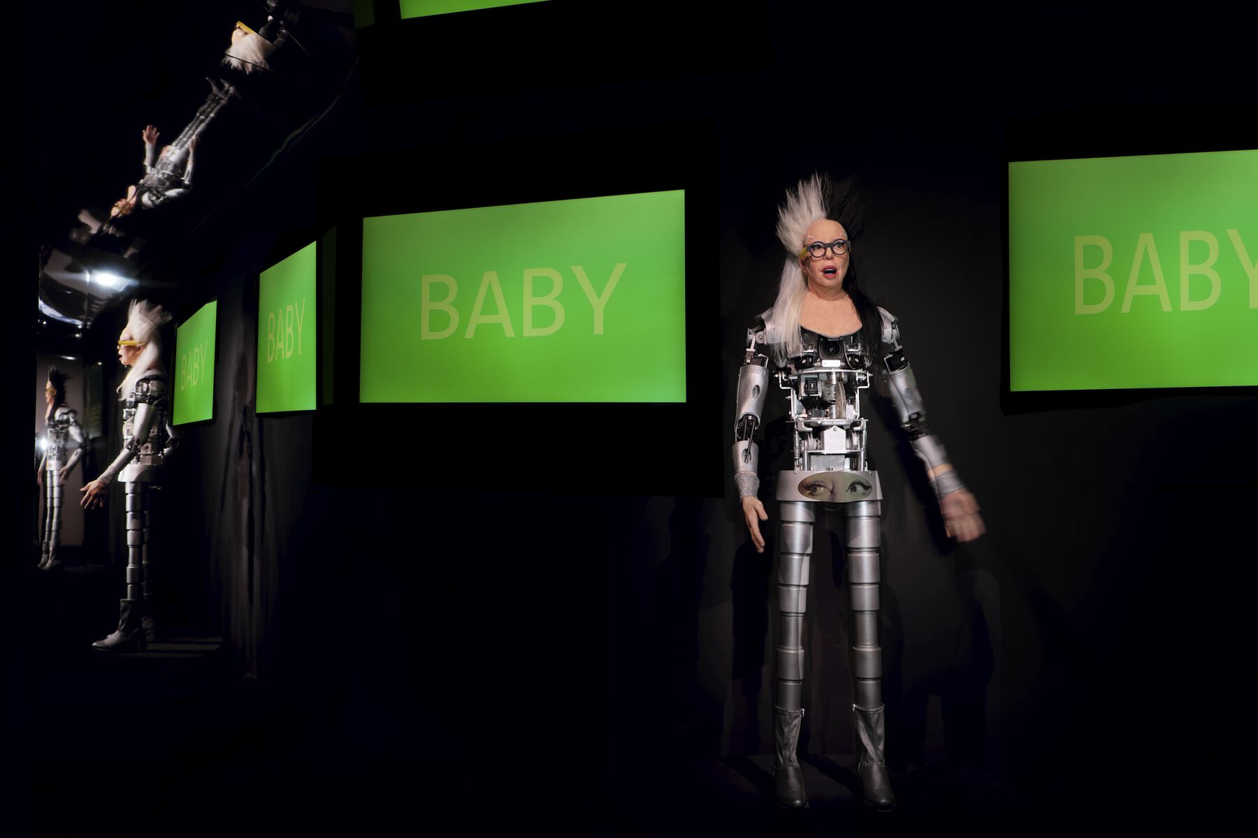 A robot with a mechanical body with the head of the artist ORLAN standing in a dark mirrored room with two T.V. screens behind it displaying green with yellow text that reads “Baby”