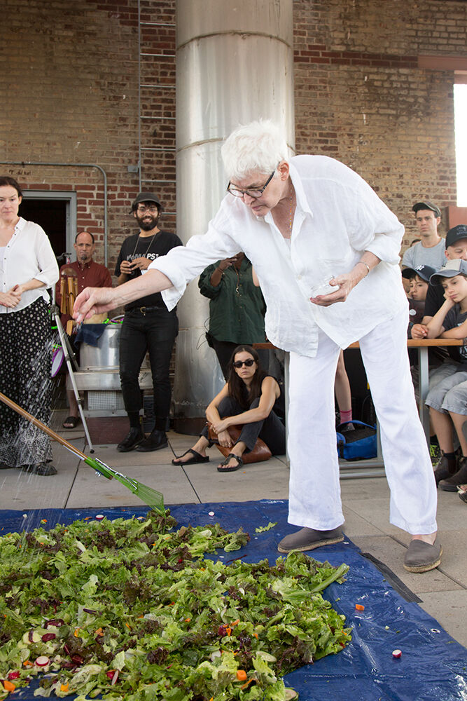 Individual dressed in all-white seasoning huge salad on blue tarp on floor as audience looks on