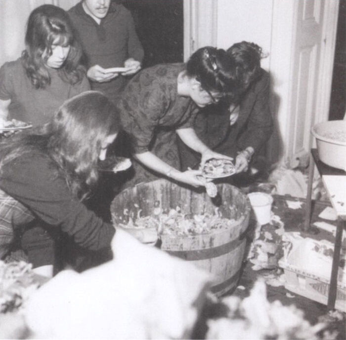 Fuzzy image of woman scooping food out of wooden barrel tub; four other figures are clustered around