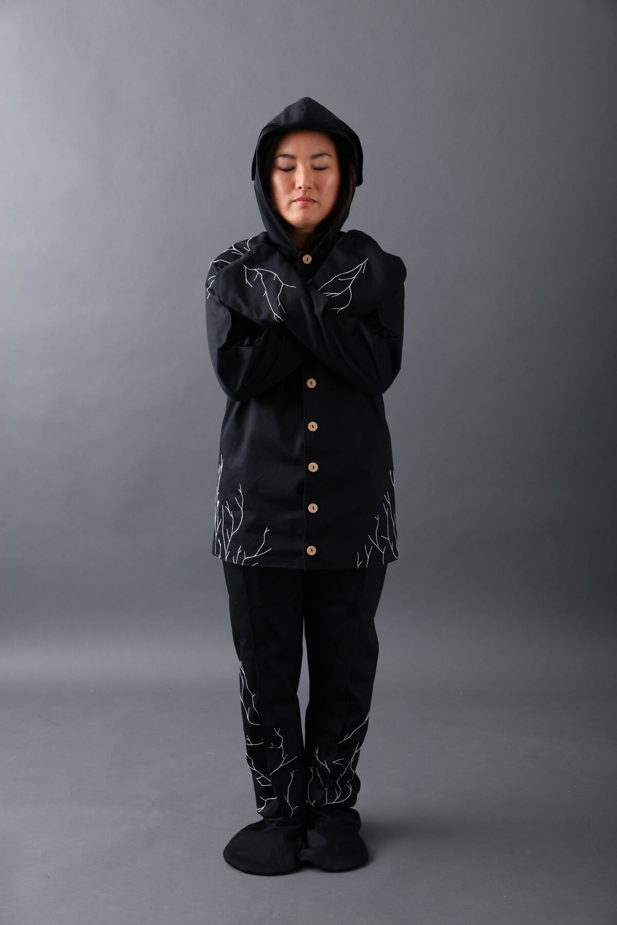 Figure in black raincoat (with hood and wooden buttons), pants, socks; veiny white embroidery on garments