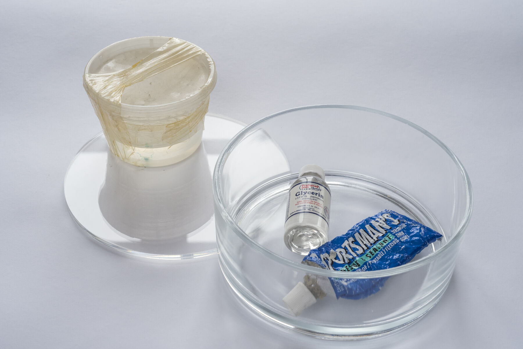 Cylindrical glass box with Glycerin and blue “Sportsman’s” sealant tube; on the side, cling-wrapped box containing green sample