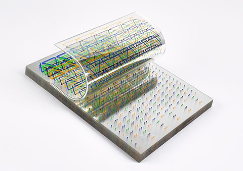 A slab of Plexiglas sheets hosts a series of green, yellow, and blue geometric patterns, with the top layer curled back in a wave formation.