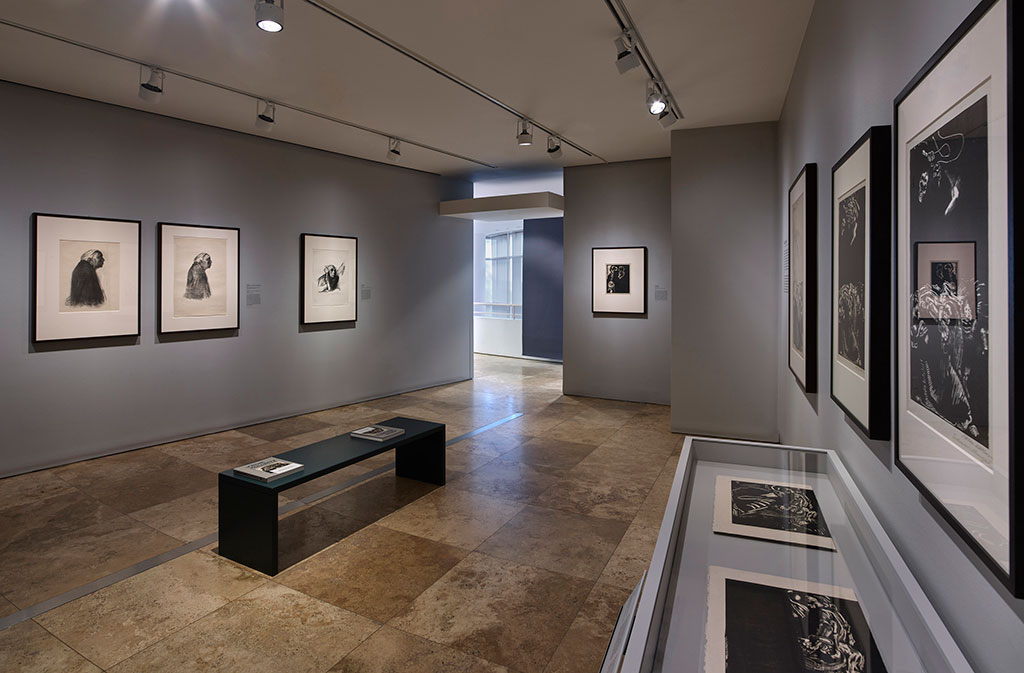 A view of the exhibition installed in gallery. Framed works line the walls and placed in a glass case. Related publications rest on a bench center of the gallery.