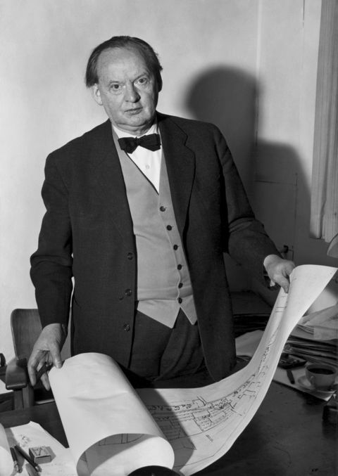 Black and white photo of Hans Scharoun dressed in a suit, standing at his desk consulting blueprints.