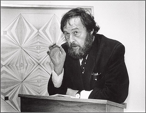 A black-and-white photograph documents the curator Harald Szeemann, a thickly bearded man with disheveled hair, standing behind a podium, delivering a lecture in front of an abstract geometric drawing by the psychic and healer Emma Kunz.