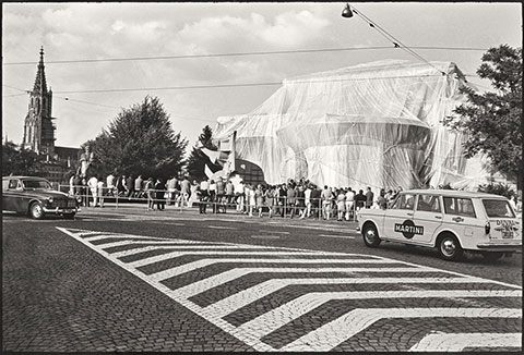 A black-and-white photograph documents a large building that has been fully wrapped in white fabric and twine, transforming it into a work of art thronged by people. The photograph was taken in the middle of a crosswalk stretching toward the building, and a taxi enters the frame on the right side.