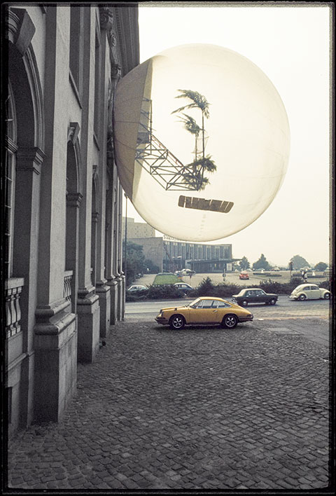 A color photograph documents an artwork by a collective of avant-garde architects: an oversized, clear bubble protruding from the side of a stone building, containing two palm trees attached to a silver gridded structure. A yellow sports car is parked on the sidewalk underneath it.