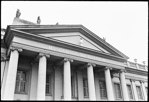 A black-and-white photograph documents a performance: the artist James Lee Byars is in a white suit, standing on the ledge of a large columned building with his back turned toward the camera.