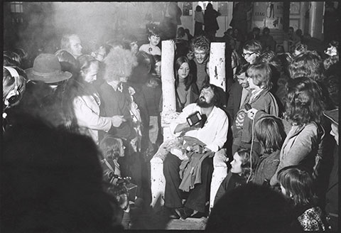 A black-and-white photograph documents the curator Harald Szeemann, a heavily bearded man in his thirties, sitting on a wooden throne in the middle of a crowd with a passport in his hand.