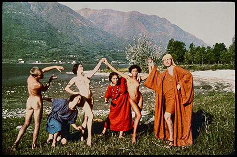 A color photograph documents a troupe of early 20th-century dancers in a grassy meadow in front of a towering mountain; three are nude and three are wearing vibrant robes as they pose with their arms raised and hands interlocking.