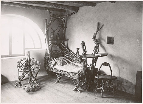 A black-and-white photograph documents a spare room with thick ceiling beams and a light-filled, half-round window. Pushed up against the wall near the window are two low chairs and a bed made from intertwined tree branches.